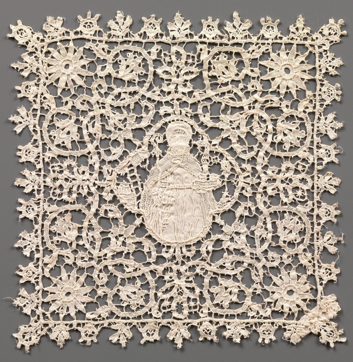 Square panel of cream needle lace worked entirely in openwork, its dense scrolling foliage surrounding a central oval medallion containing a standing bishop figure, with a deep scalloped border of points