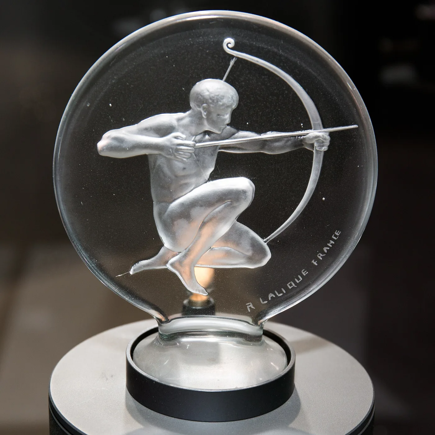 Circular glass disc enclosing a frosted archer drawing a bow, mounted on a black cylindrical base, with an engraved maker's inscription visible along the lower edge of the glass