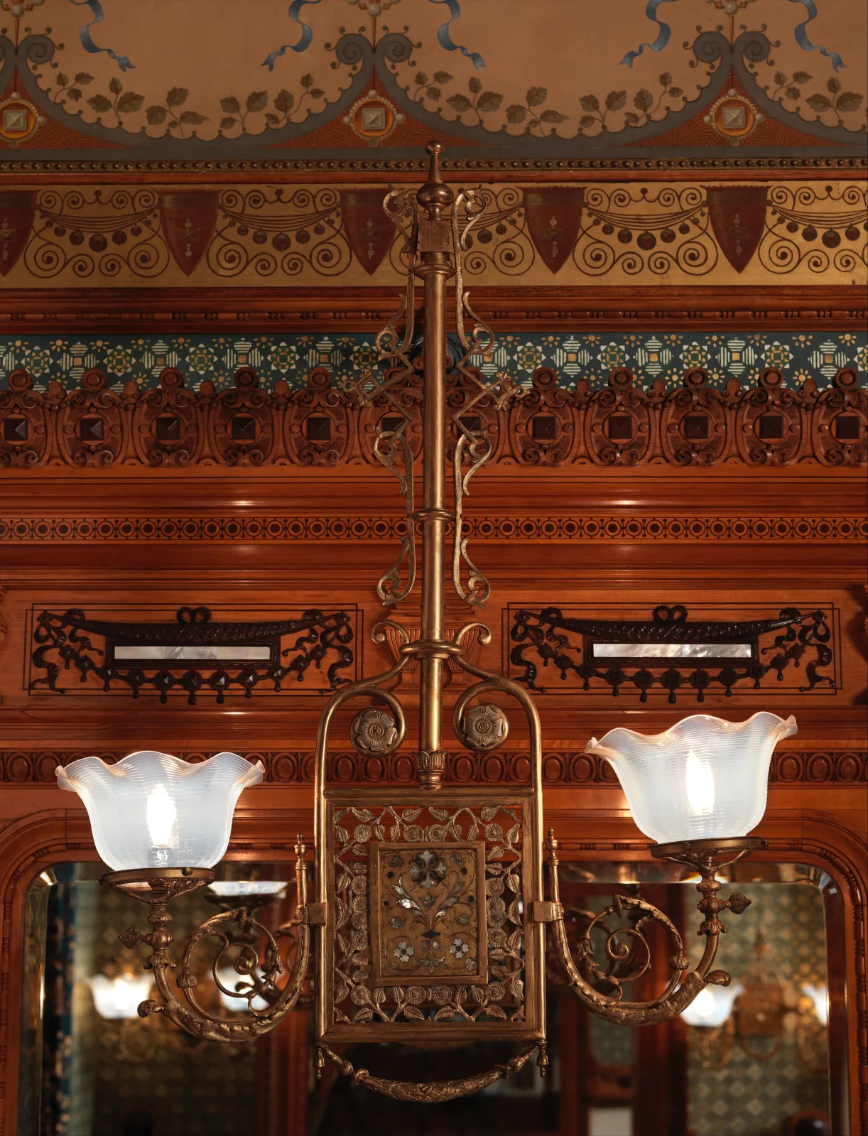 Gas wall bracket of 1881 to 82 by George A Schastey and Company, two brass arms with frilled opal glass shades flanking a pierced panel inlaid with mother of pearl, mounted on carved and inlaid woodwork with a mirror behind and the supply pipe running up the wall