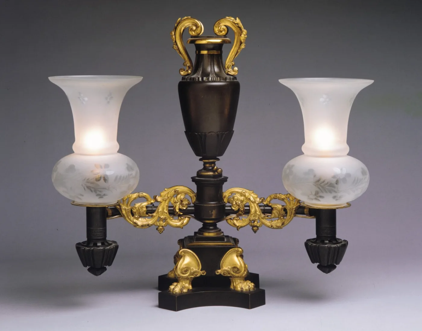 Two arm Argand lamp of 1835 to 40, a dark bronze urn shaped reservoir with gilt scroll handles raised above two gilt bronze arms that carry burners with frosted etched glass shades, on a triangular plinth with gilt paw feet