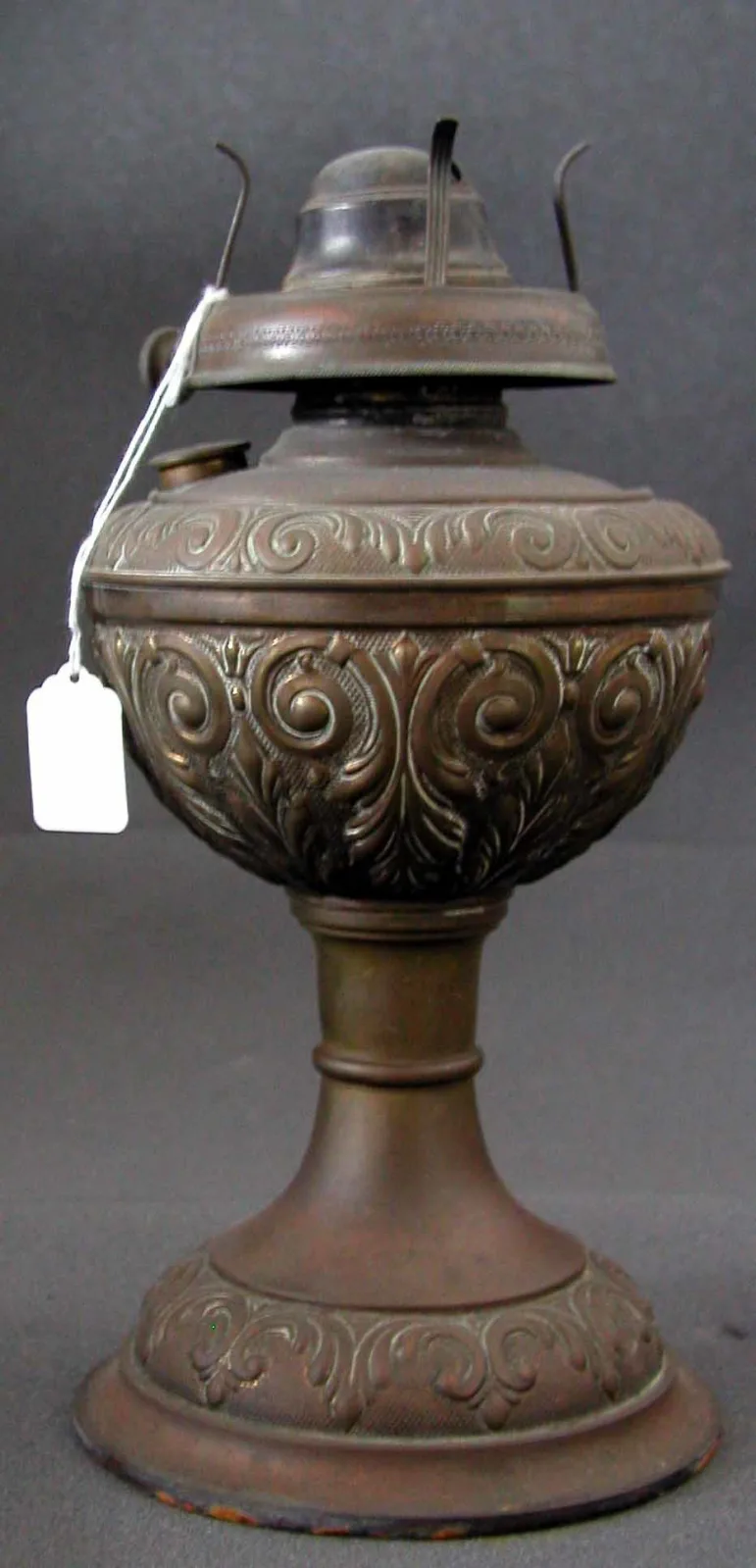 Kerosene lamp with an embossed brass font of scrollwork in two registers on a matching decorated foot, fitted with a burner and chimney holder prongs but no chimney or shade