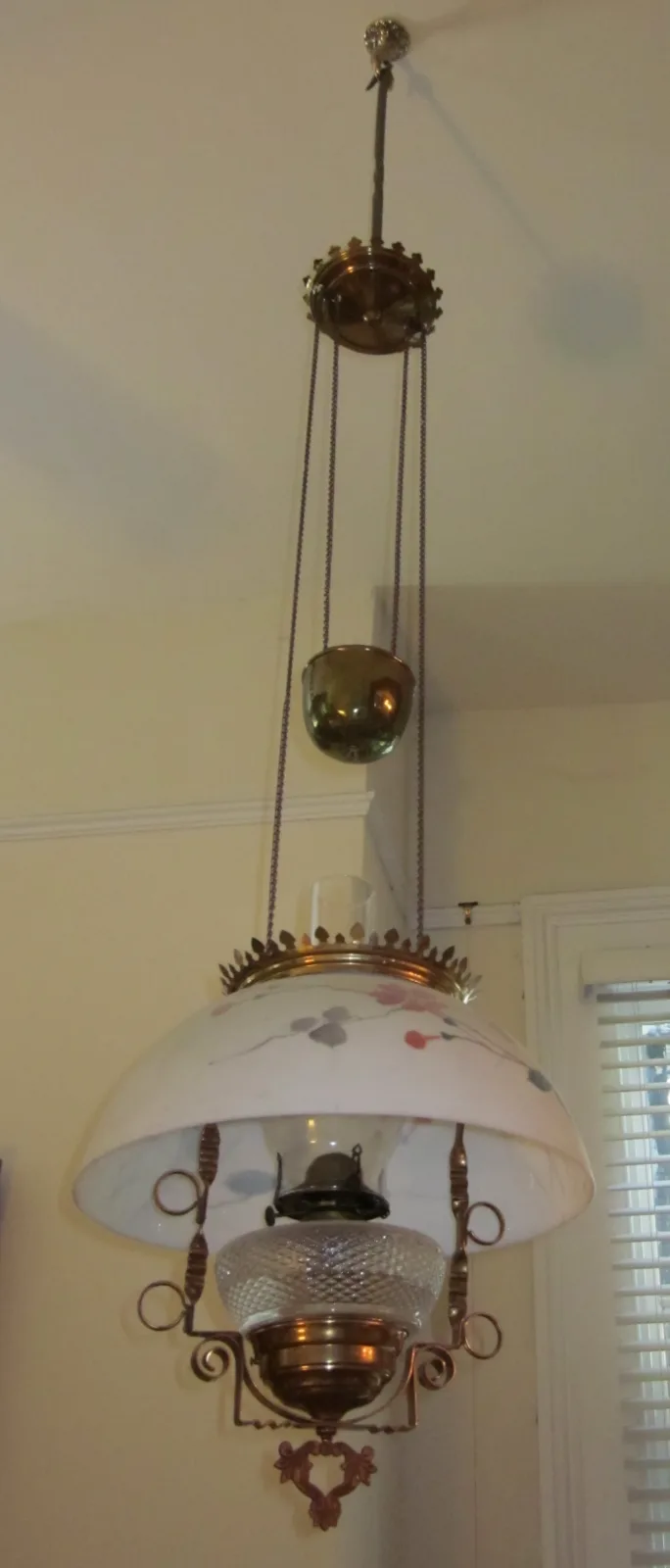 Pendant oil lamp hanging on a chain from a ceiling rose with a pulley and counterweight above, a white glass dome shade below, and the lamp font with burner beneath the shade