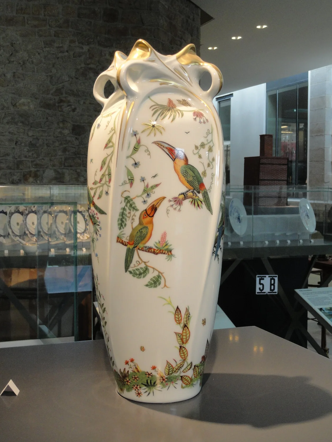 Tall white porcelain vase with looping gilded handles growing from the shoulder, painted with toucans among tropical foliage