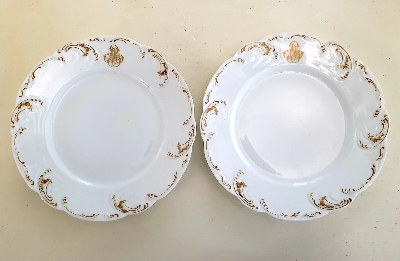 Two white porcelain plates with moulded rococo scrollwork rims, a gilt monogram at the top and gilding worn away to dull patches