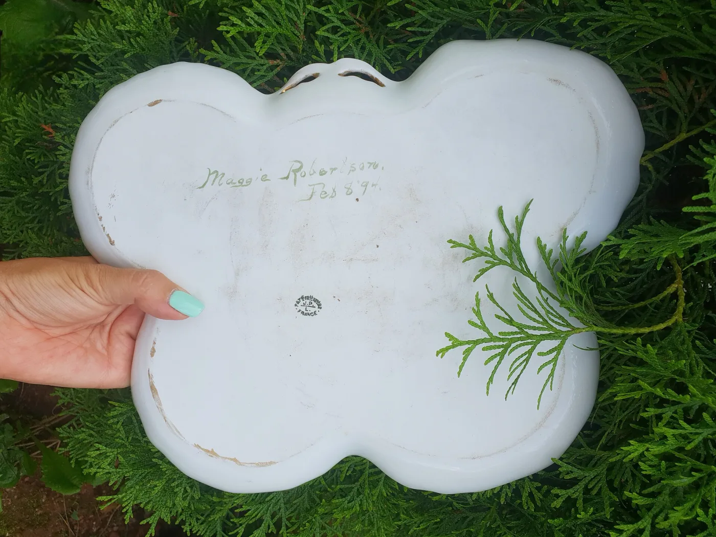 Underside of a butterfly-shaped white porcelain tray held in one hand, showing a green painted signature reading Maggie Robertson Feb 8 94 above a small green oval factory stamp