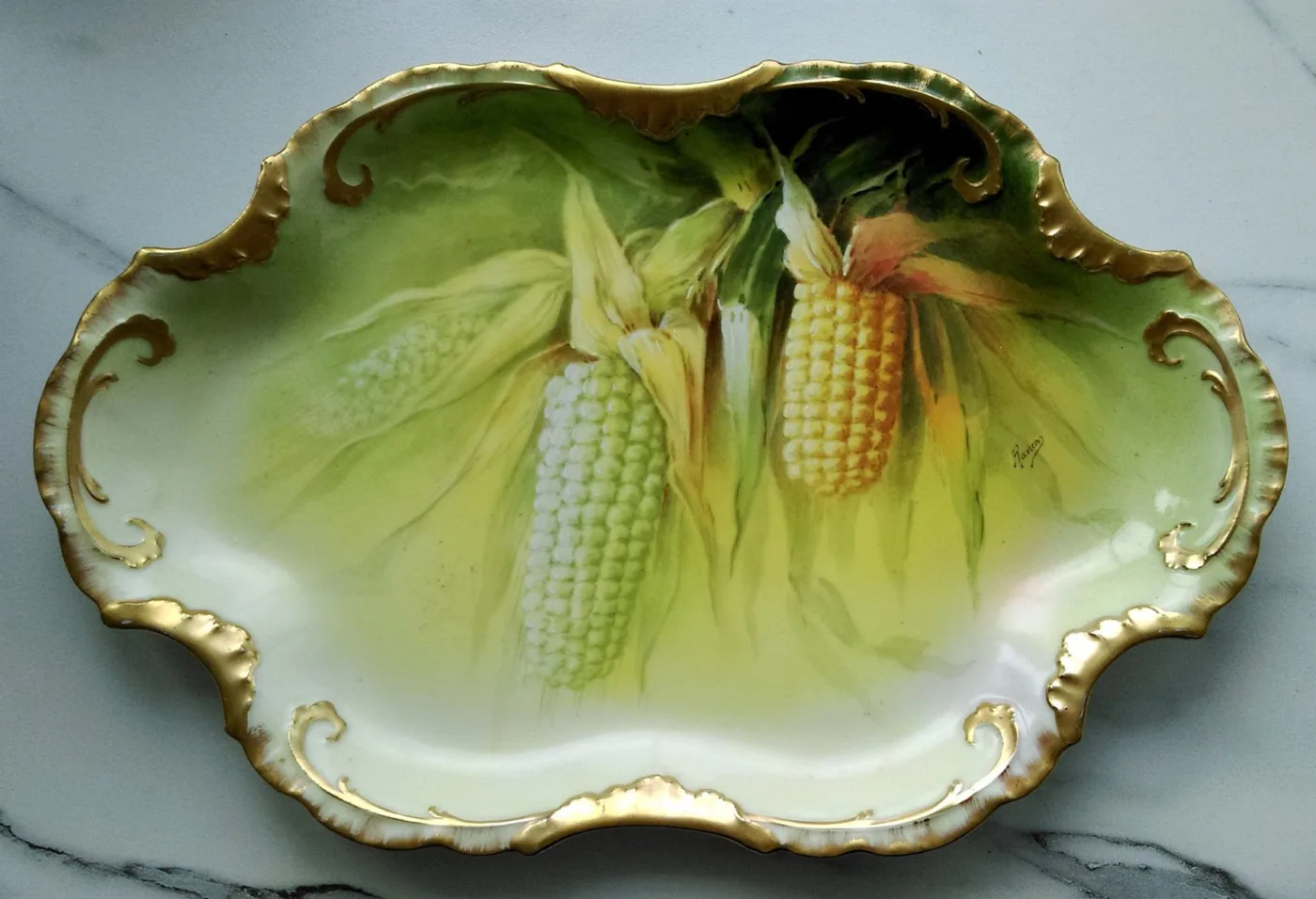 Green and gold porcelain tray hand-painted with ears of corn in their husks, with a small painted signature reading Rancon at the right edge