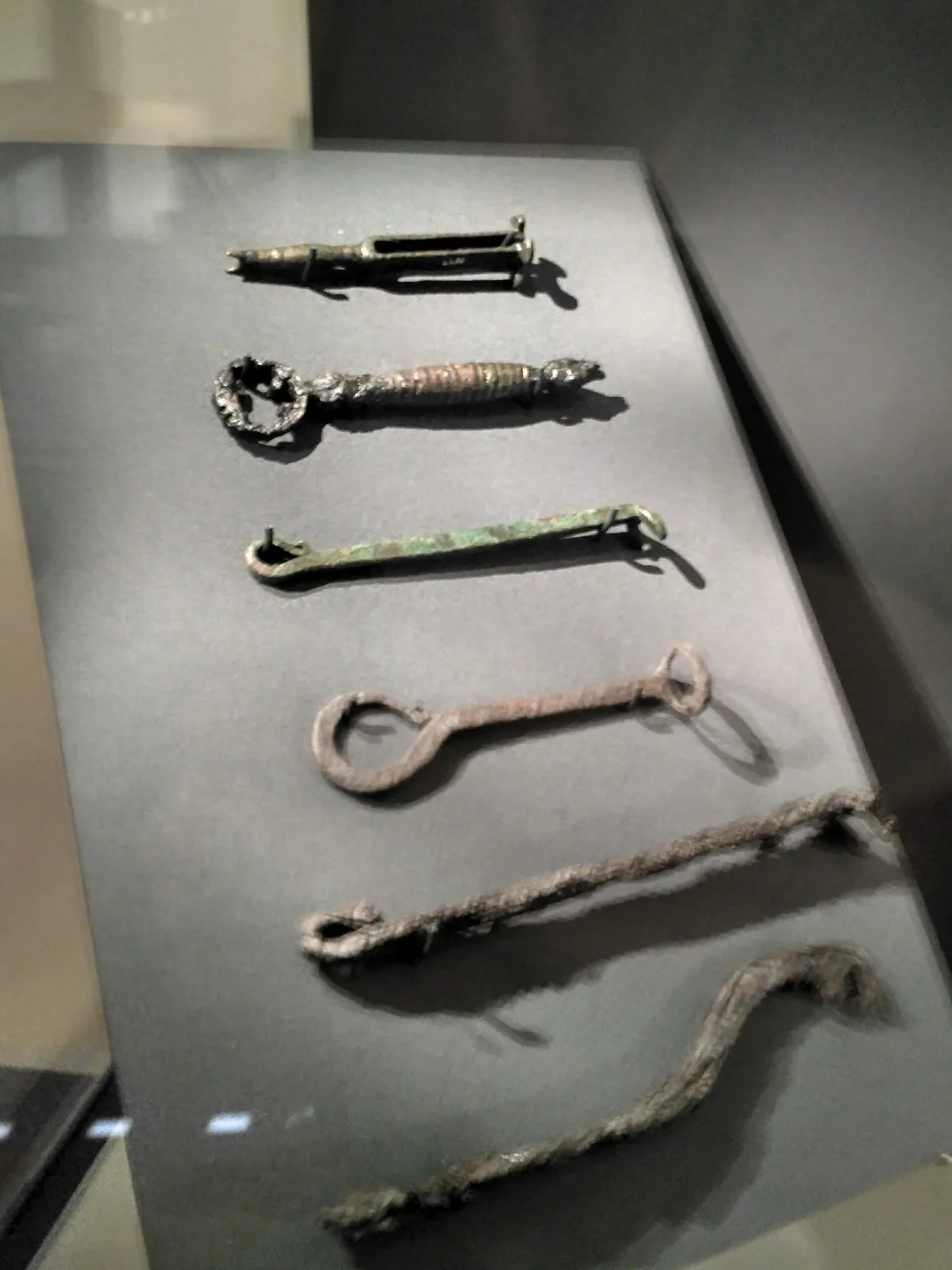 Six corroded iron and copper-alloy objects of different forms displayed on an angled grey museum mount: a slender barrel mechanism, a key with an openwork ring bow and ribbed shank, a flat pierced strip, a key with a large ring head, and two long twisted rods with looped ends
