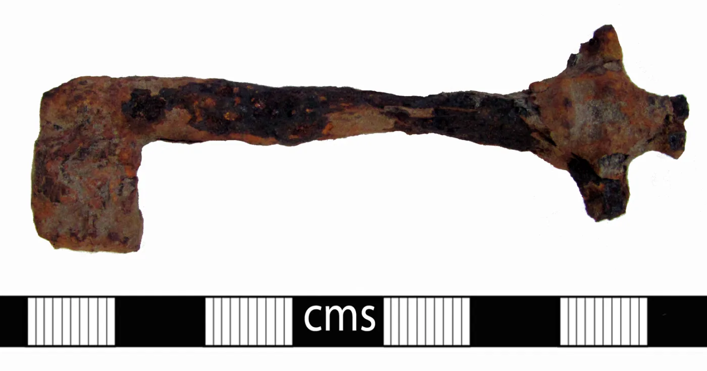 Heavily corroded iron key laid against a centimetre scale, its surface swollen into rough orange-brown crust, the head missing and the bit reduced to an indistinct lump
