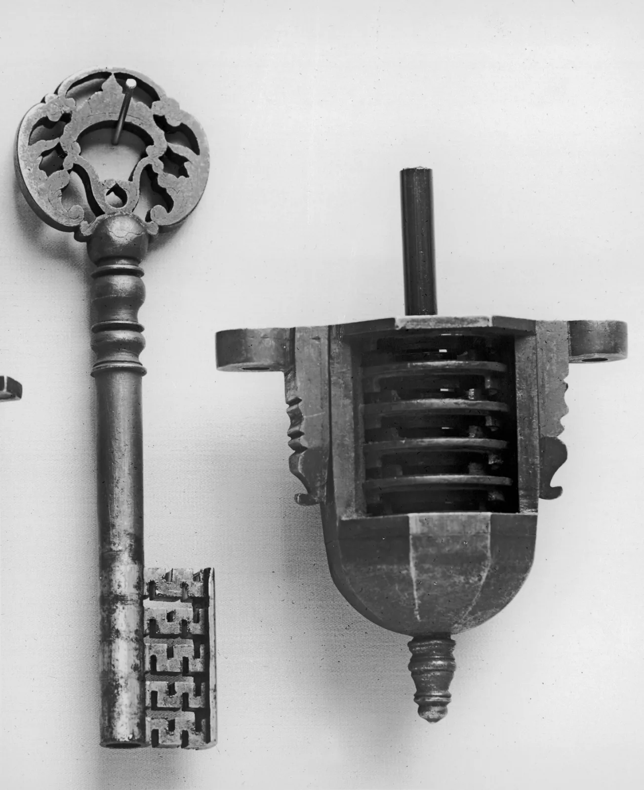 Ornate iron key with a pierced and scrolled bow and a complex stepped bit, photographed in black and white beside the lock nut it operates, a small block fitting with a projecting threaded bolt