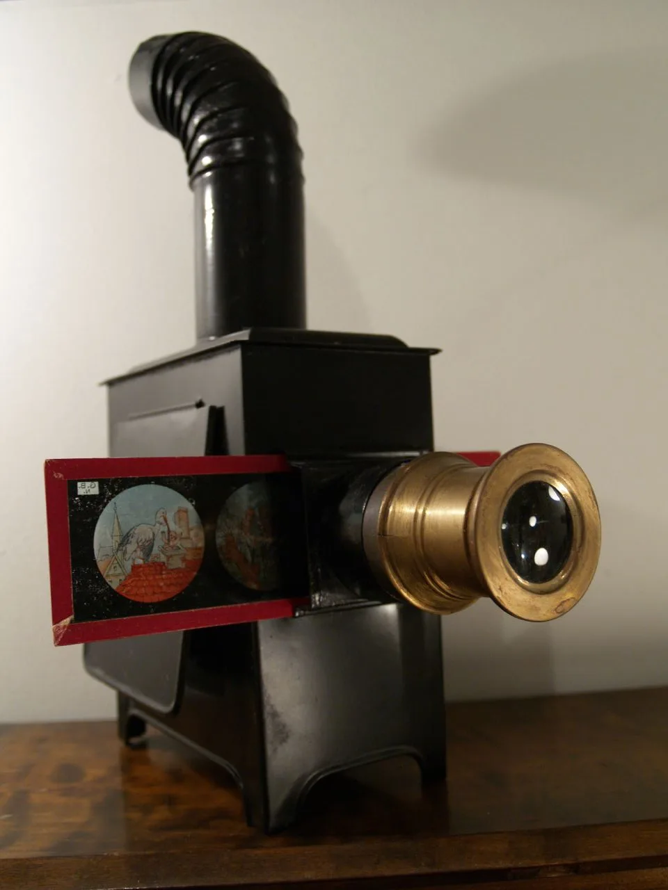 Black sheet-metal magic lantern with a tall jointed chimney rising from its top, a brass lens tube projecting from the front, and a red-framed glass slide pushed into a slot behind the lens