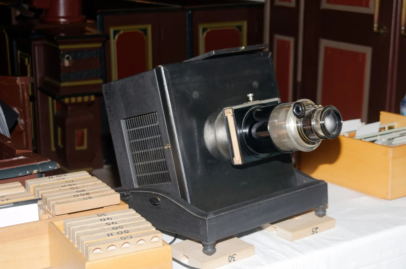 Large black lantern with a long lens barrel and a stepped body standing on a table, with stacked wooden boxes of slides ranged behind it in a panelled room