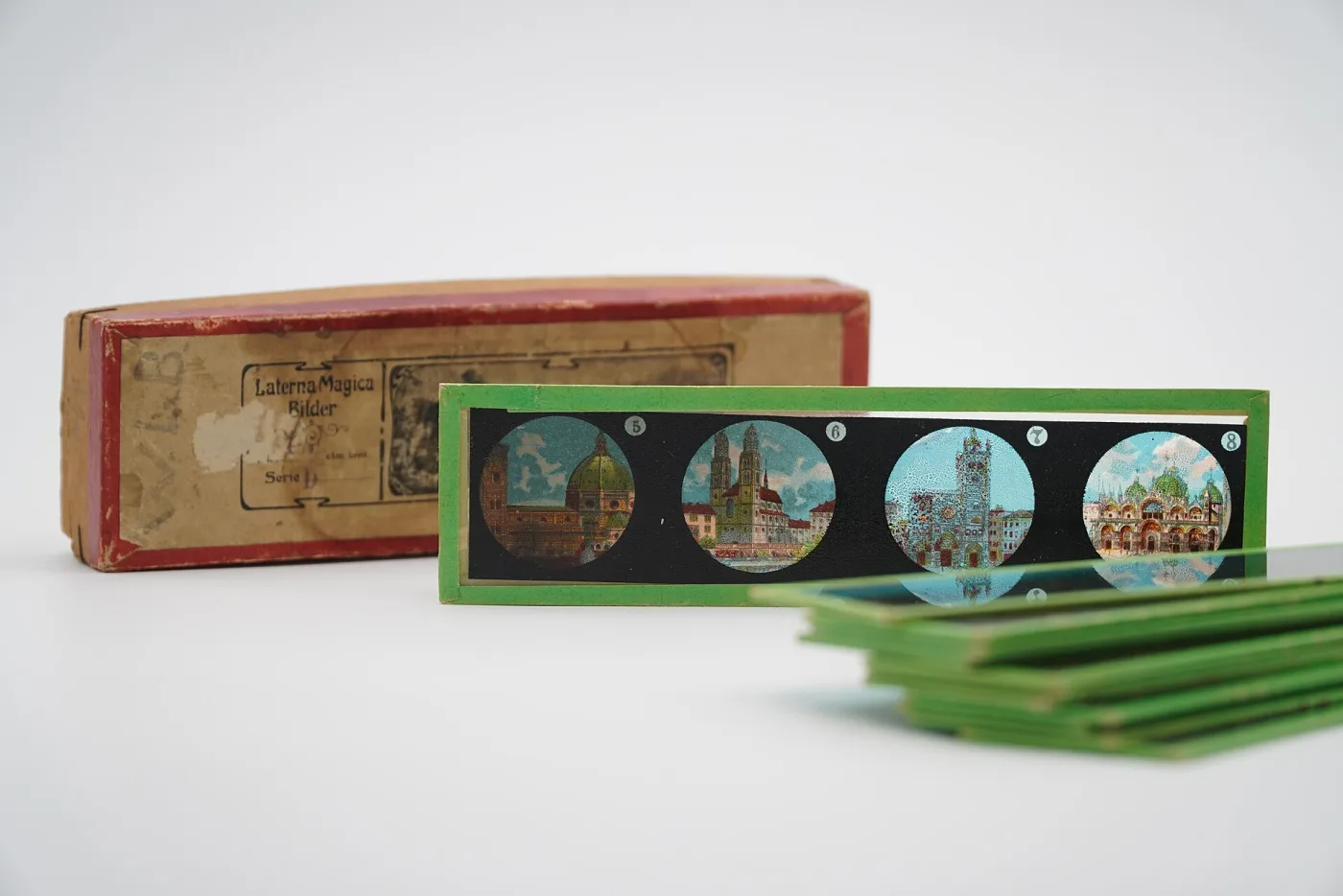 Long shallow printed cardboard box lying open with a fan of green-framed lantern slides drawn out of it, each carrying four small circular images in a row