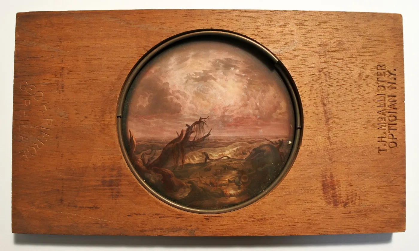Rectangular mahogany slide frame with a circular window at its centre holding a hand-painted glass image of a dark mountainous landscape under a heavy sky