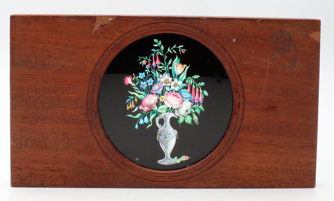 Reddish mahogany slide frame with a circular window holding a hand-painted image of a bouquet of flowers in a classical vase against a black ground