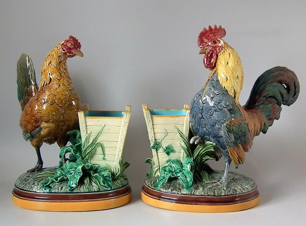 Pair of majolica vases modelled as a hen and a rooster standing beside cylindrical fluted holders, on oval bases with green foliage
