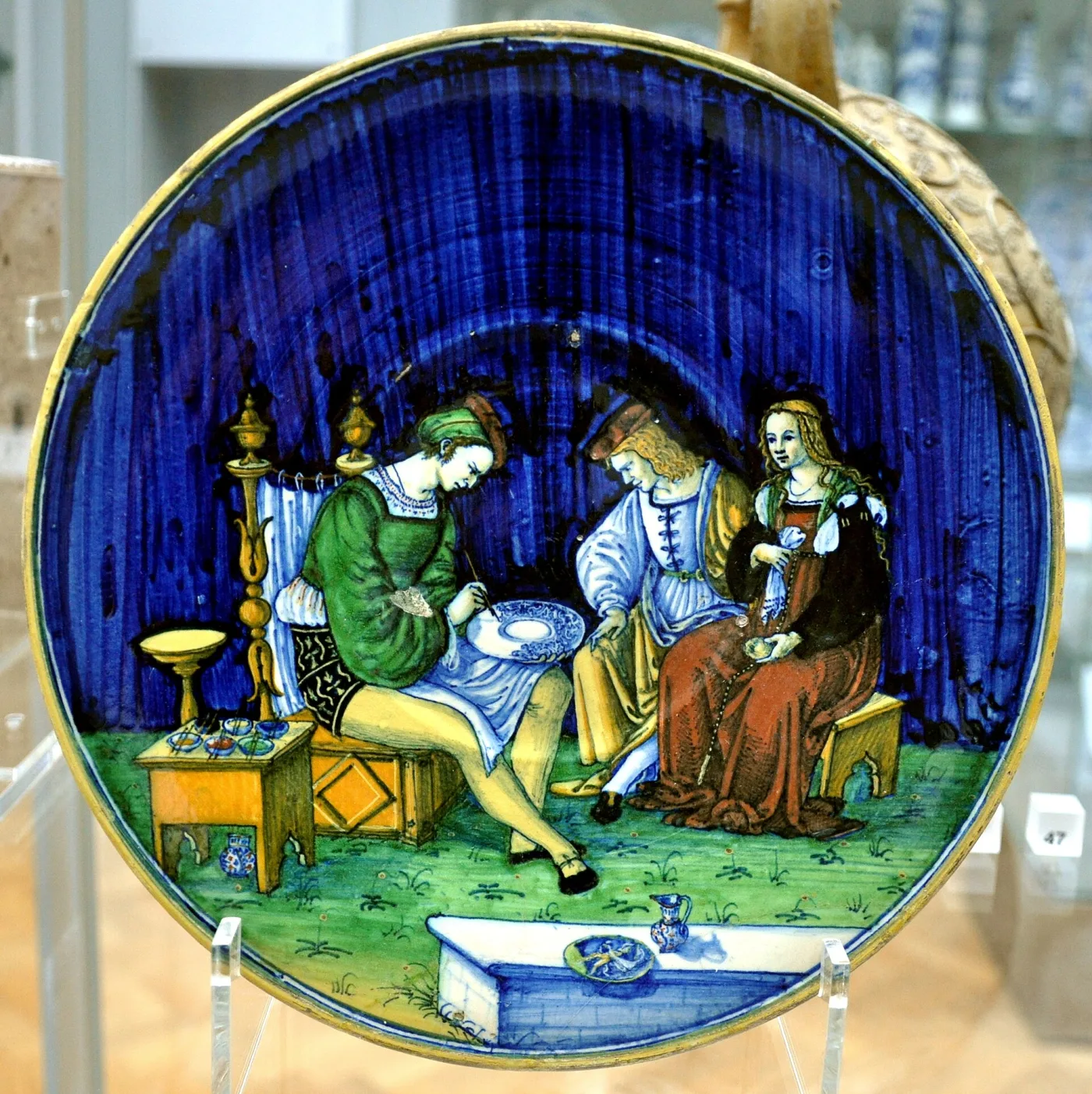 Dish with a deep blue ground showing a maiolica painter at work at a table decorating a dish, watched by three figures