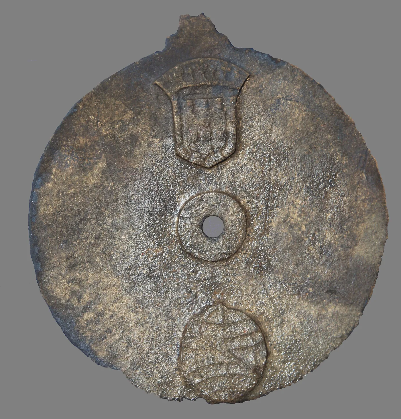 Heavily corroded disc of dark metal with a pitted, encrusted surface, bearing a raised royal shield at the top and a second armillary device below, with a small central hole