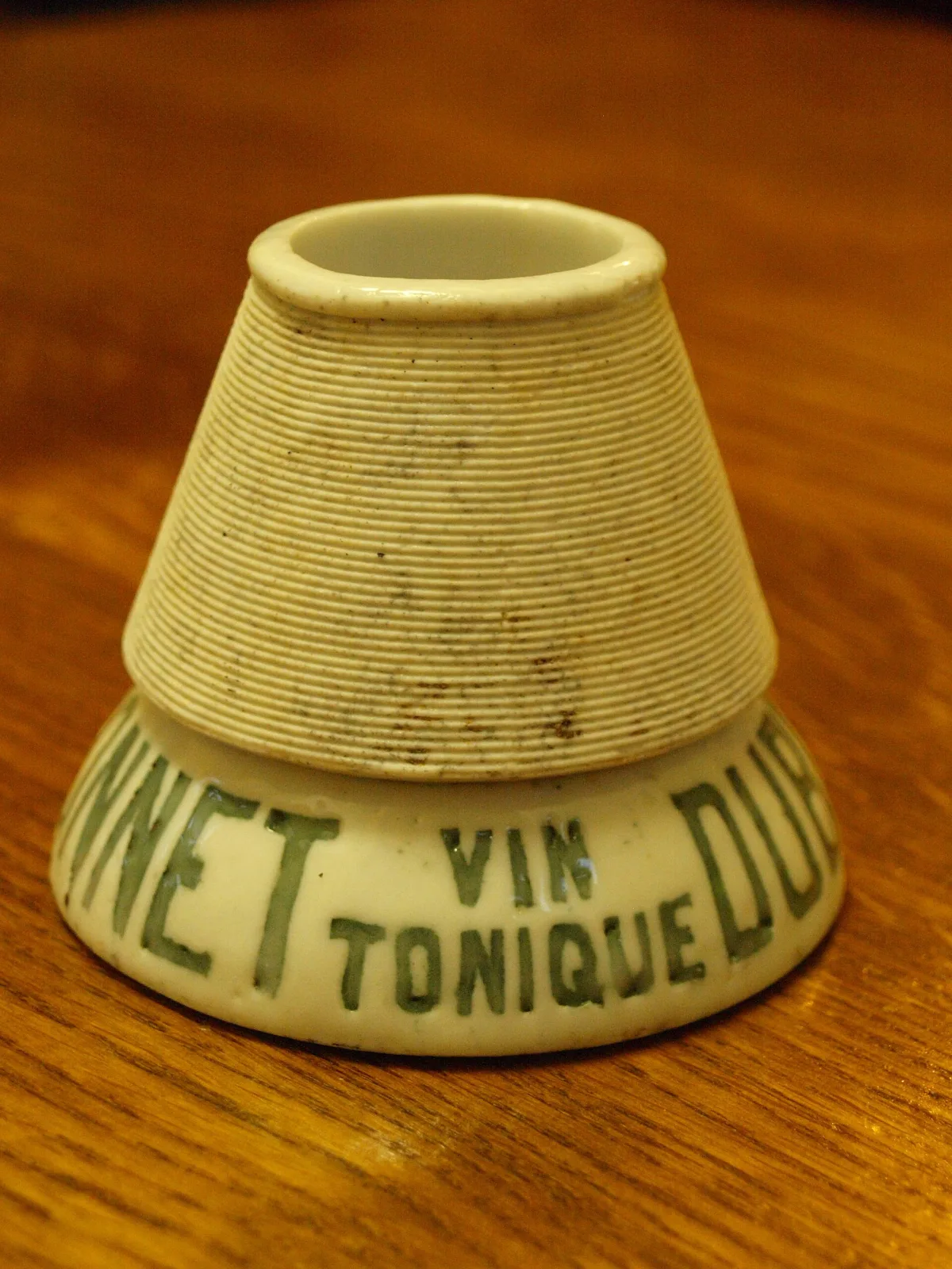 Conical white porcelain match striker, its cone finely ribbed all over and stained brown from use, standing on a flared base lettered in green with a wine advertisement