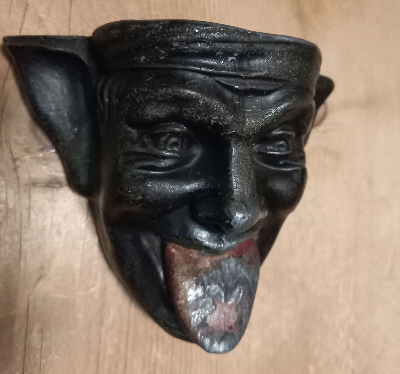 Cast iron wall plaque in the form of a horned devil&rsquo;s head with pointed ears, its long red tongue protruding and worn bare down the centre
