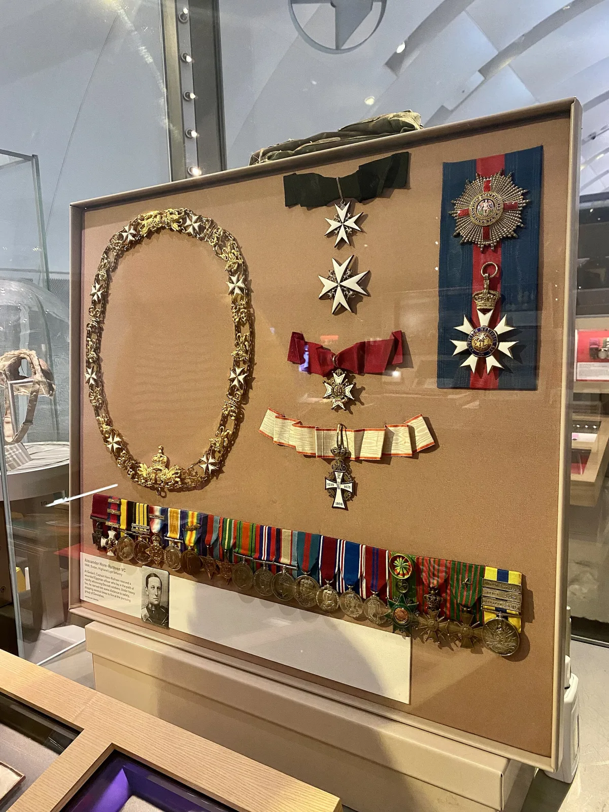 Museum display case containing a gold collar chain, several enamelled neck badges on ribbons, two breast stars and a long mounted row of miniature medals, arranged on a fawn board