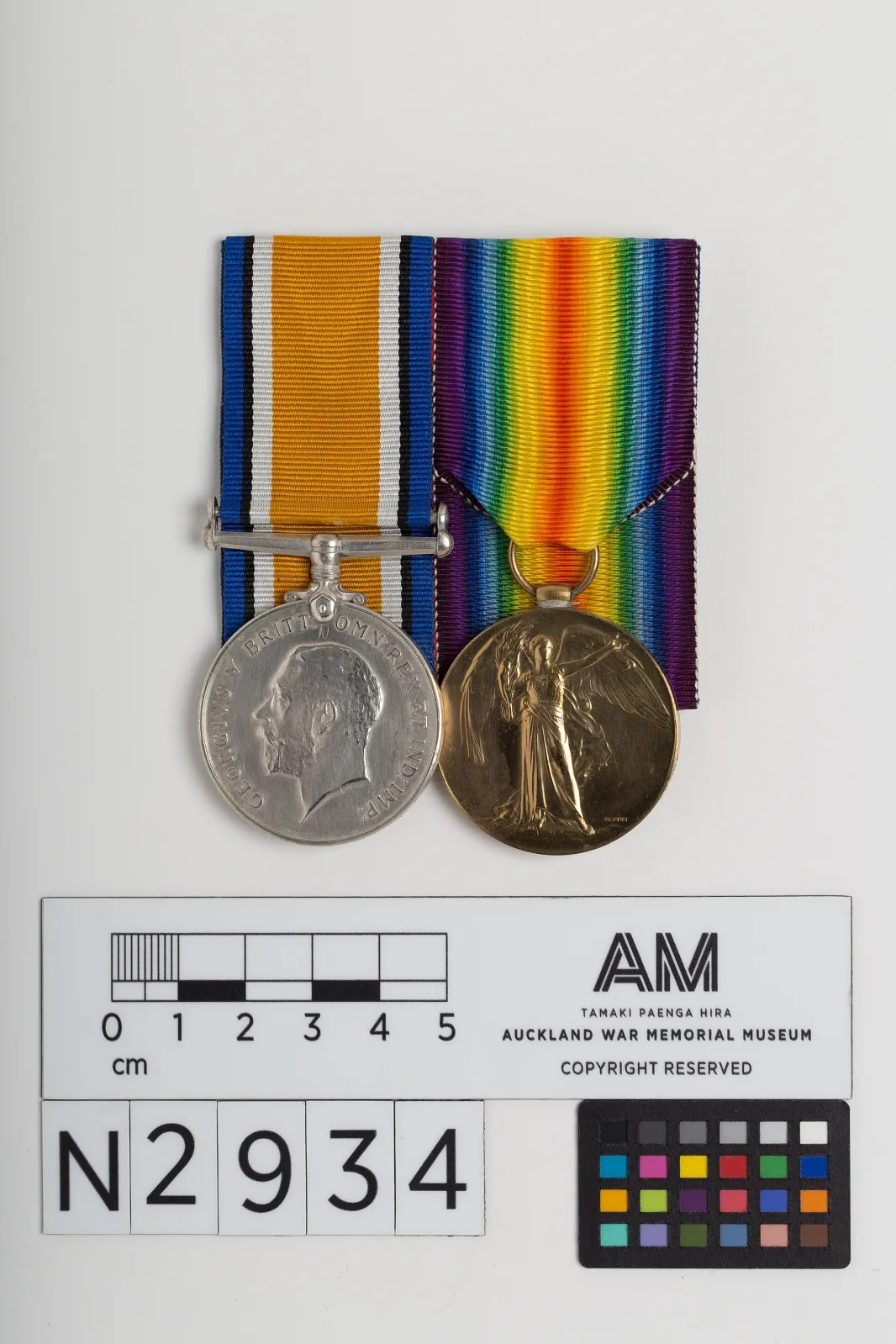 A pair of First World War medals on their ribbons beside a museum scale bar and colour chart printed with the accession number N2934, one silver with a bare-headed king's effigy, one bronze with a winged figure