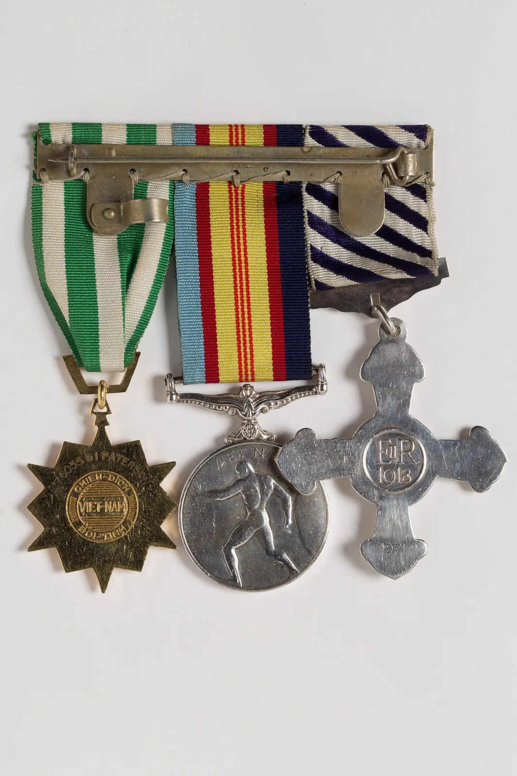 The reverse of a mounted group of three medals, showing the long pin brooch bar they hang from, the folded ribbon ends stitched over it, and the plain reverses of the cross, medal and star