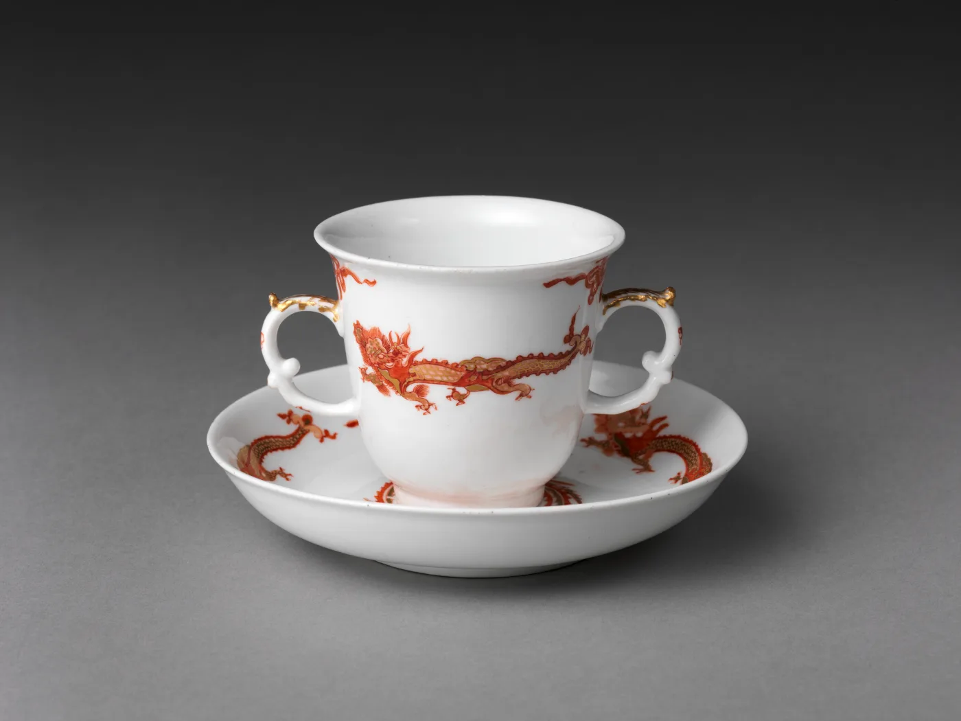 Two-handled porcelain chocolate cup and its saucer, painted in iron red and gold with slender dragons and long-tailed phoenixes among scrollwork on a white ground