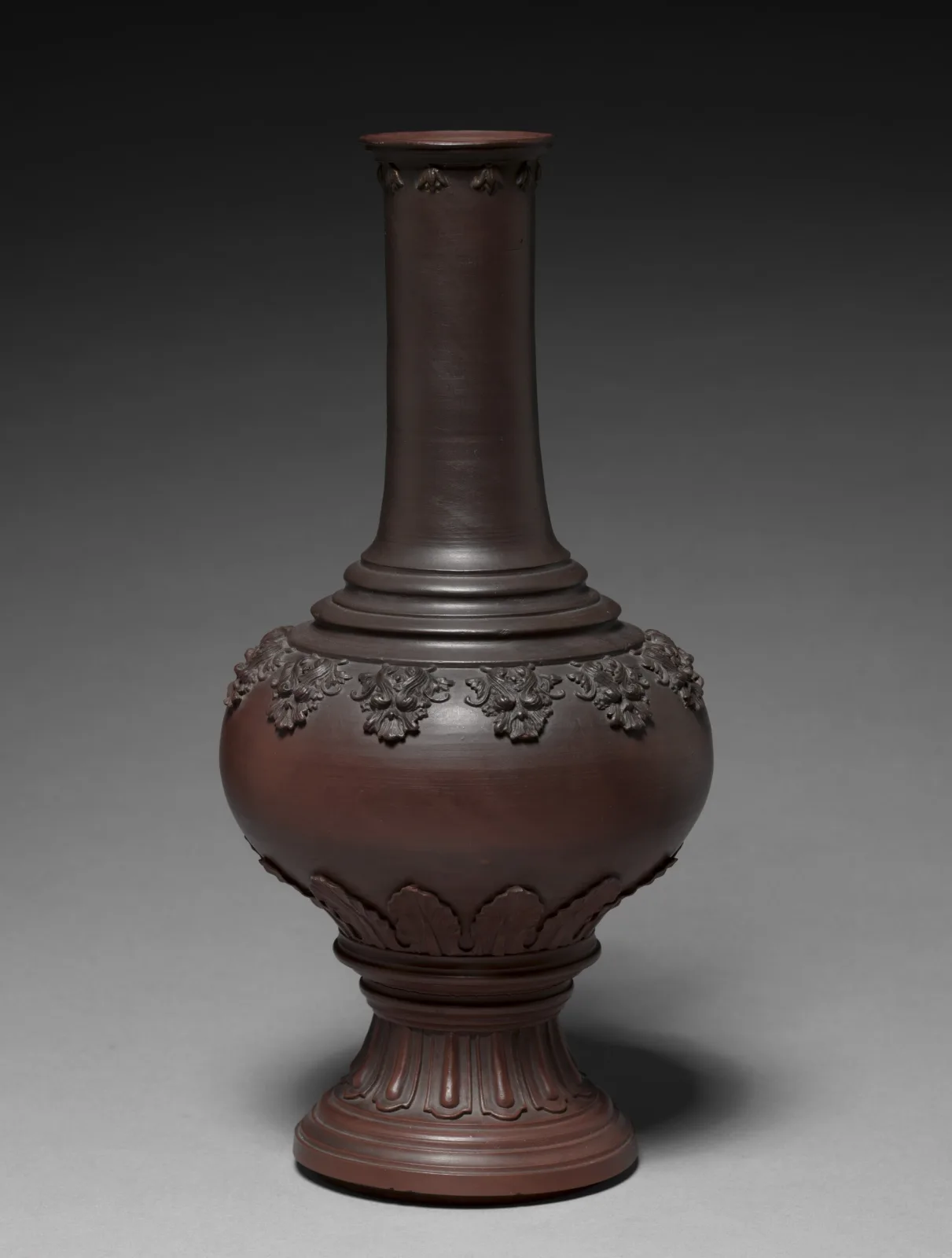 Tall bottle-shaped vase in dense red-brown stoneware, unglazed and matt, with a moulded band of ornament round the shoulder and a stepped moulded foot