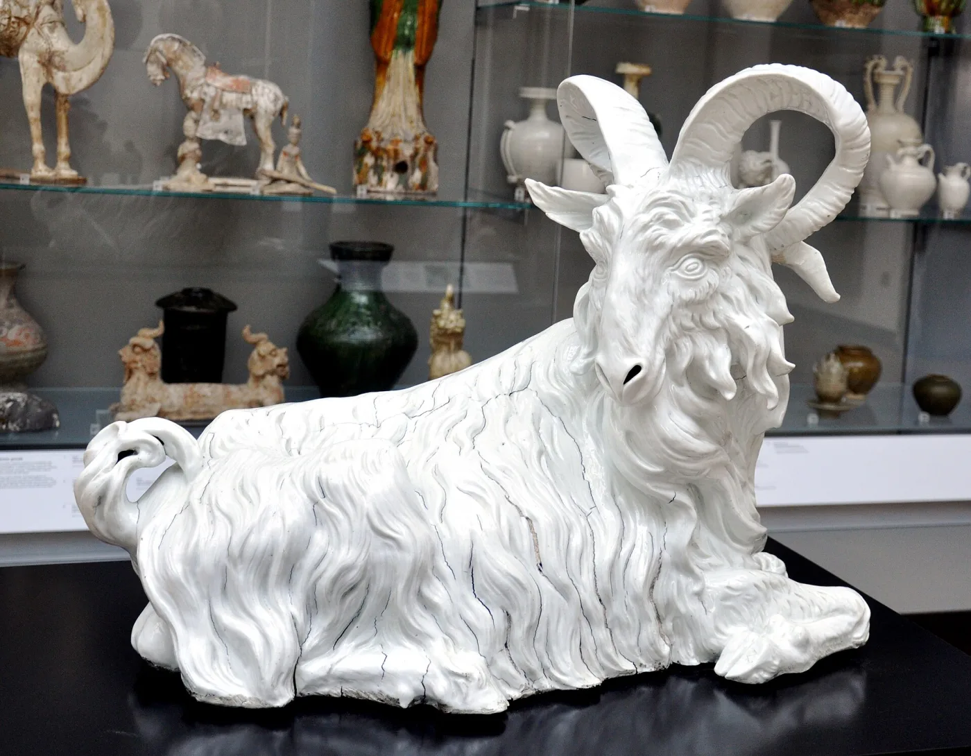 Large white porcelain goat lying with its legs folded beneath it, modelled with deeply cut shaggy hair, curling horns and a beard, undecorated so that only the modelling reads