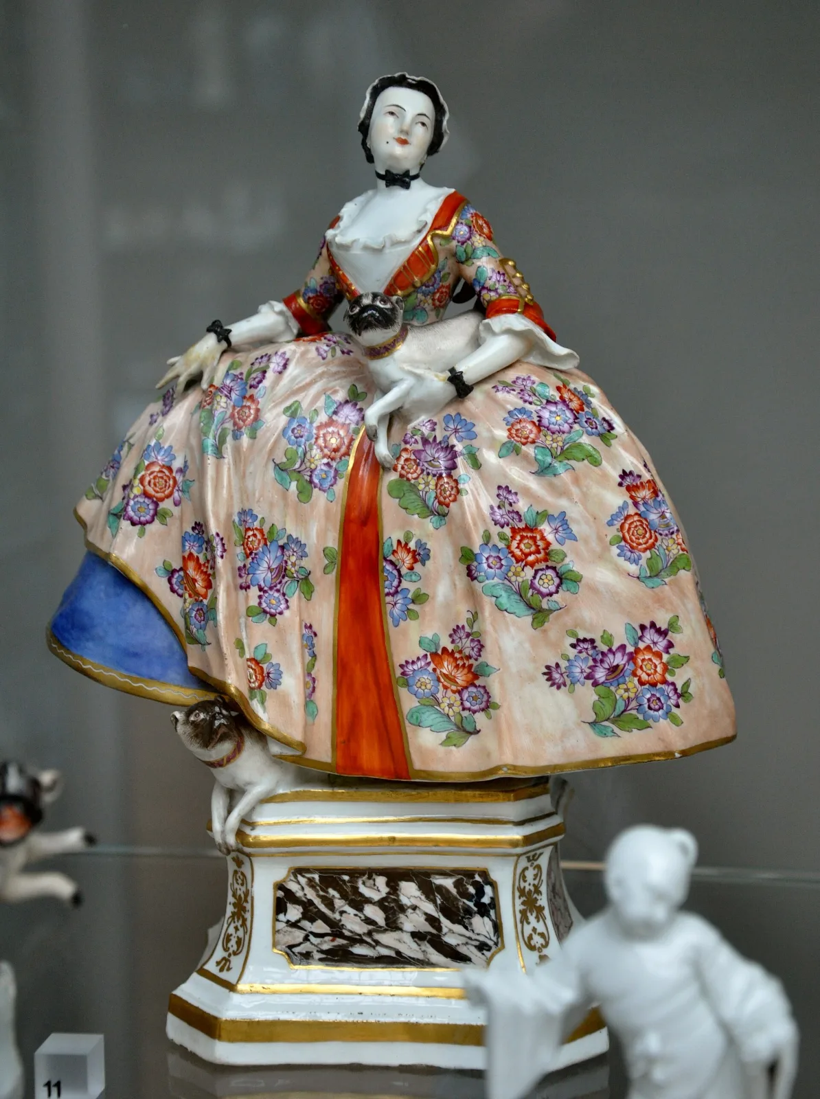 Porcelain figure of a lady in a wide flowered crinoline gown seated on a gilt and marbled octagonal plinth, one pug held in her lap and a second climbing the base beside her skirt