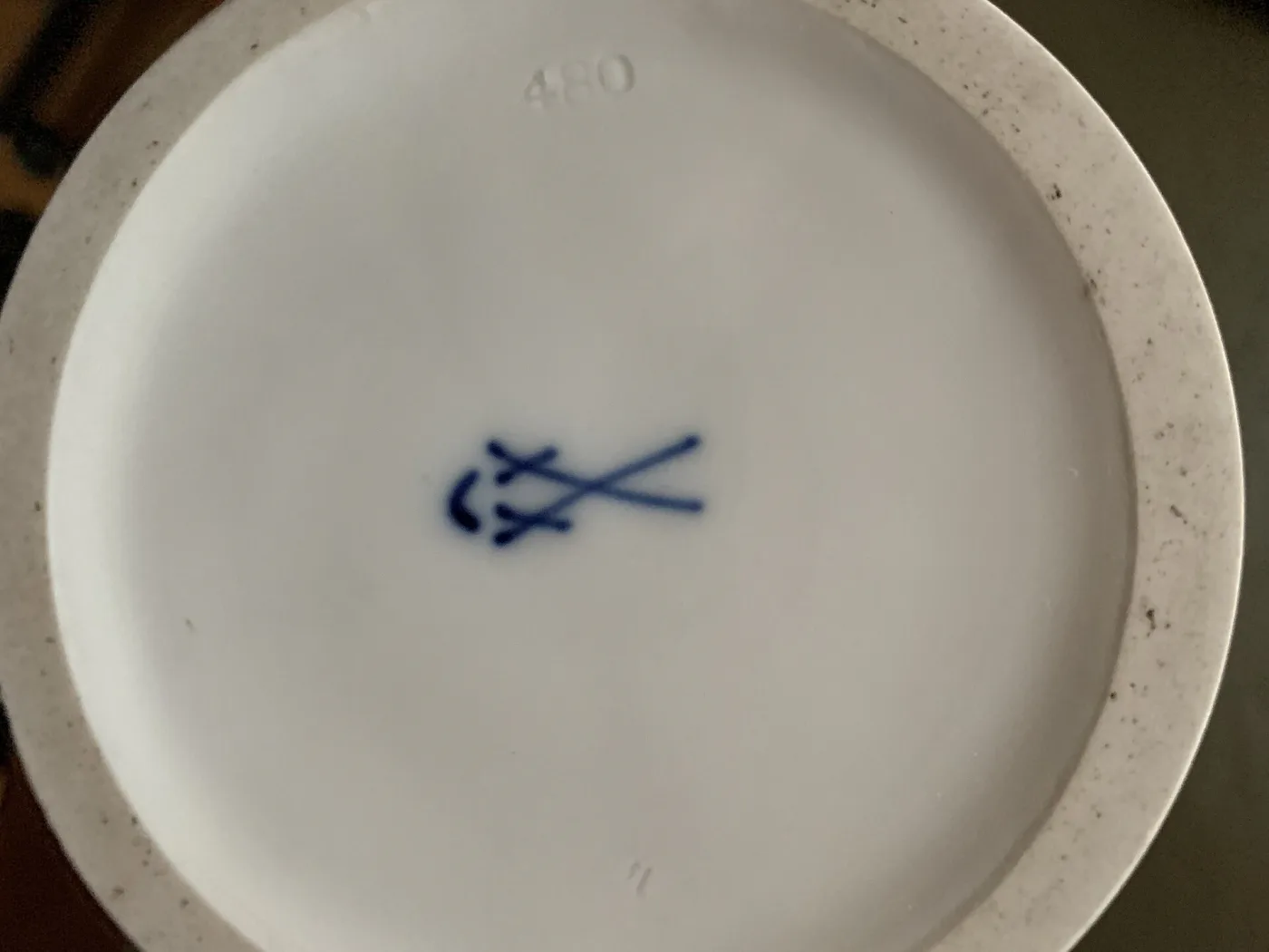 Underside of a white porcelain foot ring showing a crossed-swords mark painted in underglaze blue at the centre, with the number 480 impressed into the paste above it and a further impressed numeral below