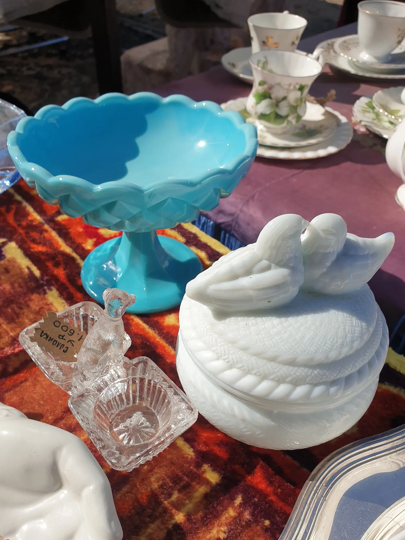 Turquoise opaque glass compote beside a white milk glass covered dish topped with two doves, on a flea-market table