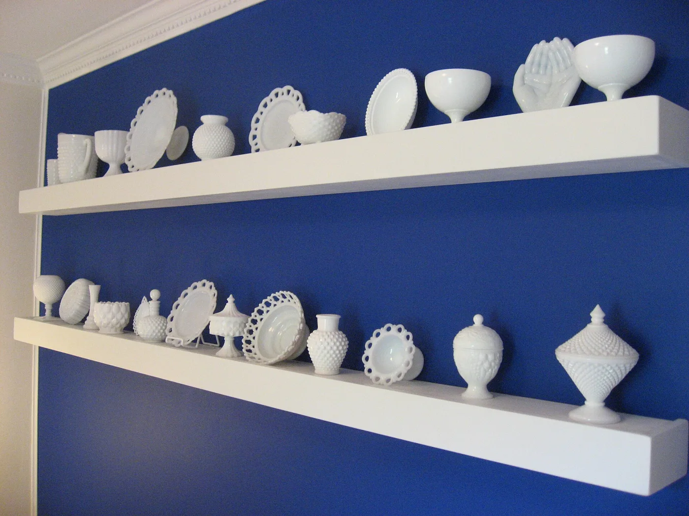 White milk glass vases, compotes and covered dishes arranged along white shelves against a deep blue wall
