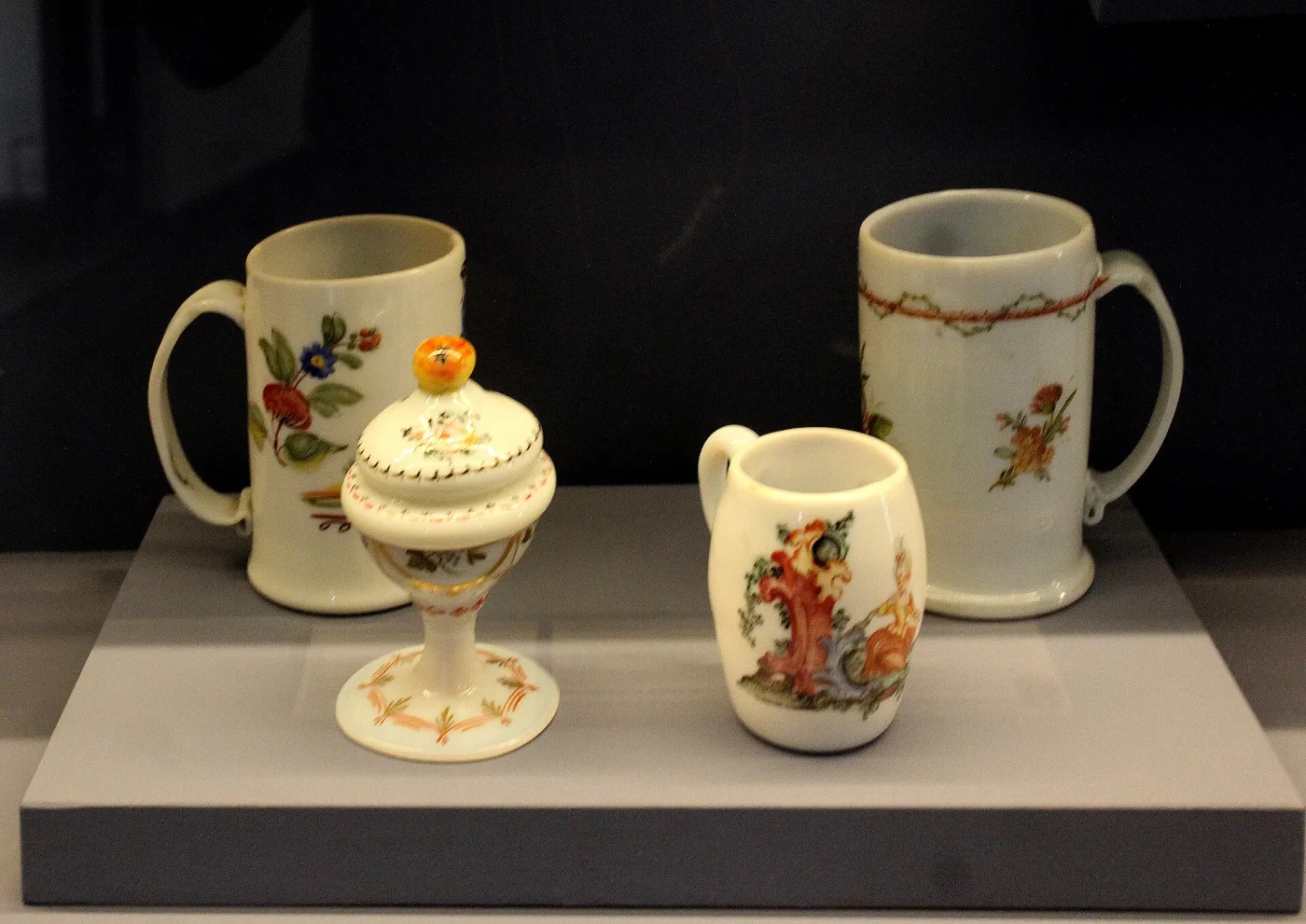 Four white tankards and a covered goblet painted with flowers, displayed on a plinth in a museum case