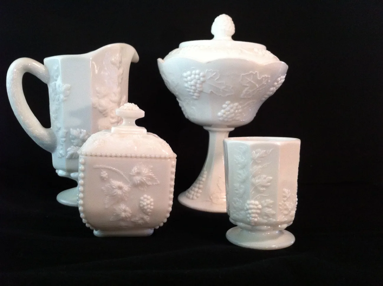 Four white milk glass pieces moulded with grape clusters and leaves — a pitcher, a covered box, a footed covered compote and a tumbler — on a black background