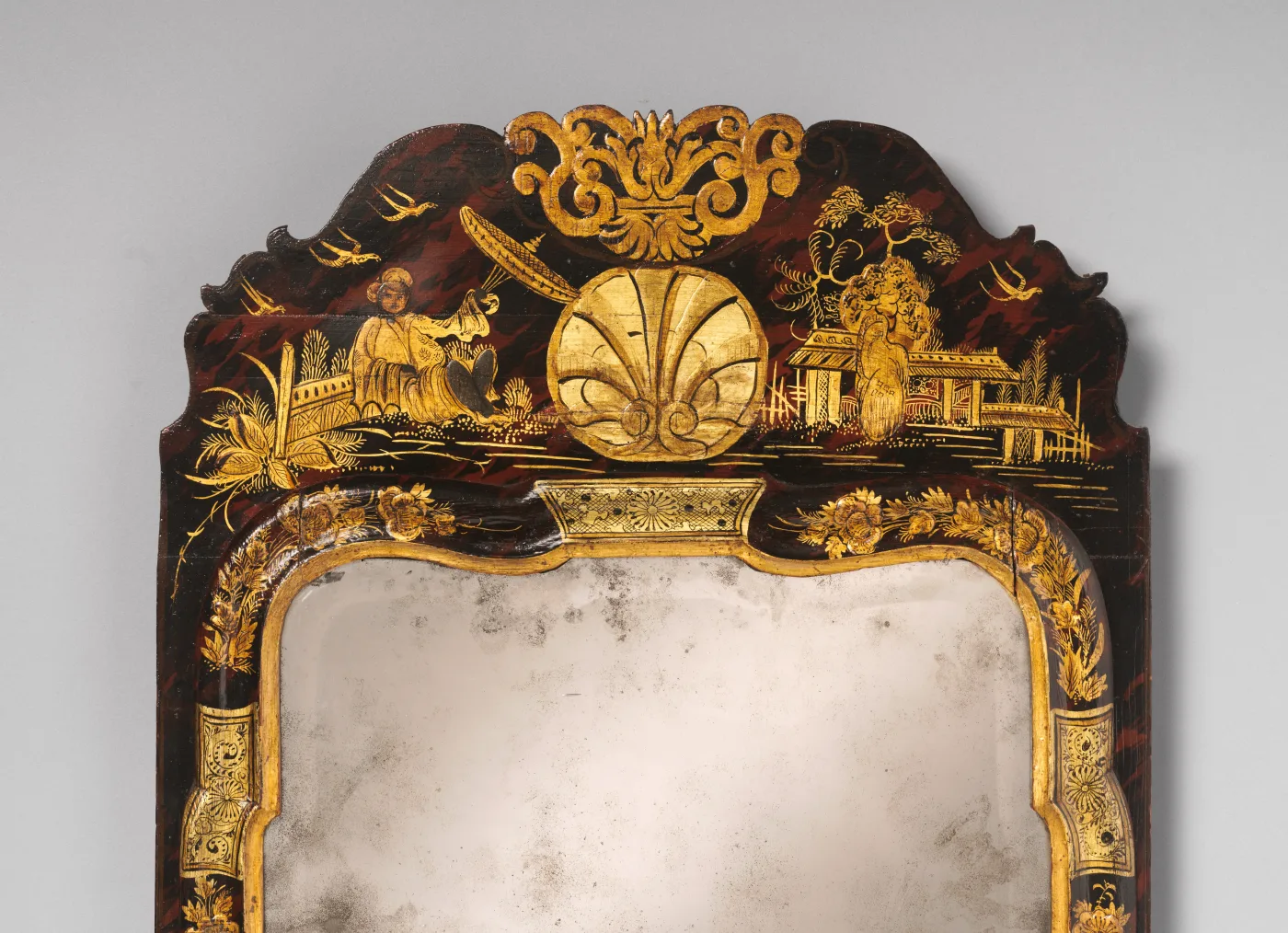 Close view of the arched top of a japanned looking glass with a gilt shell and figures on a dark ground, and below it the upper glass plate clouded with grey speckles and patches