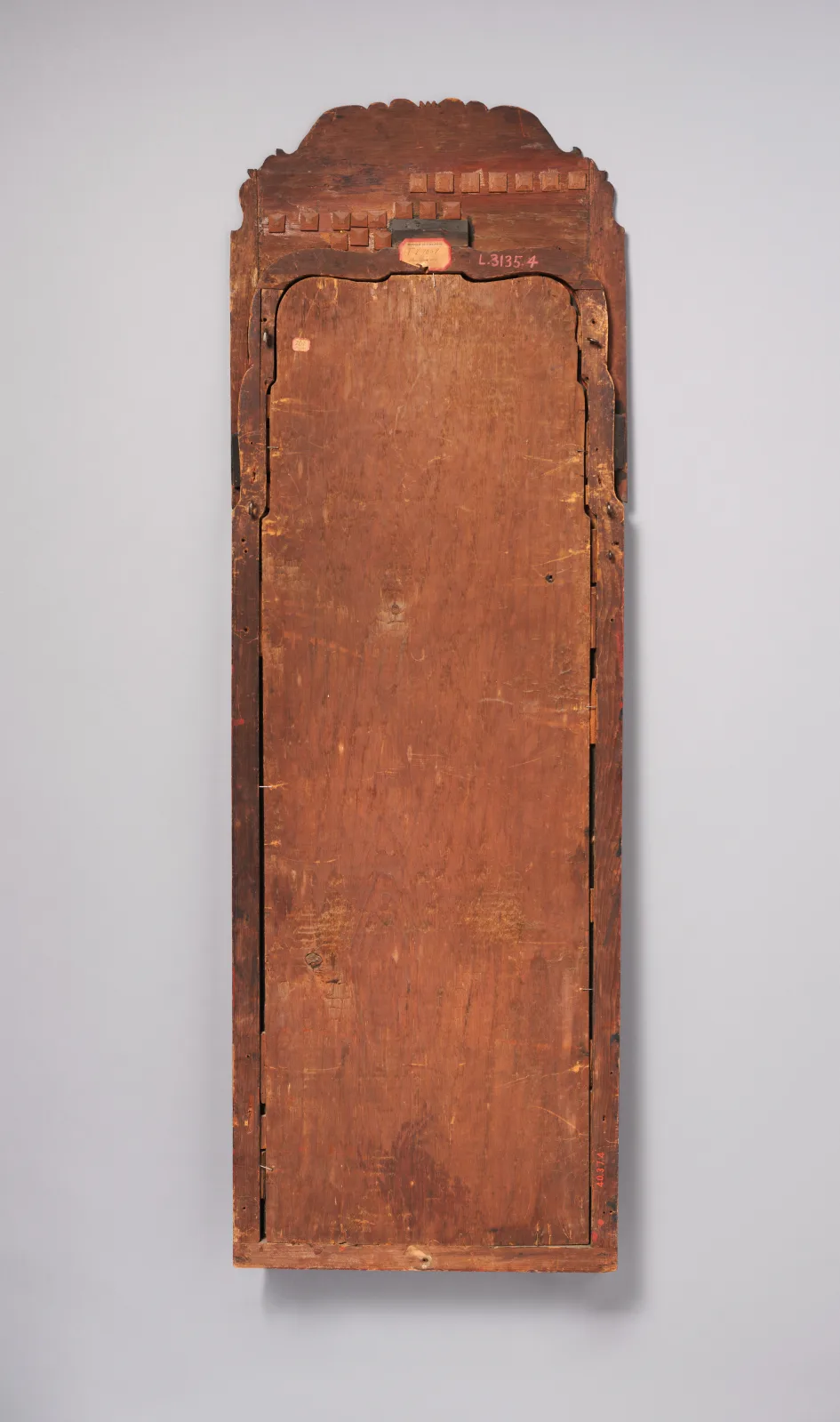 Back of a tall narrow looking glass showing plain brown wooden backboards inside the frame, with the shaped crest at the top