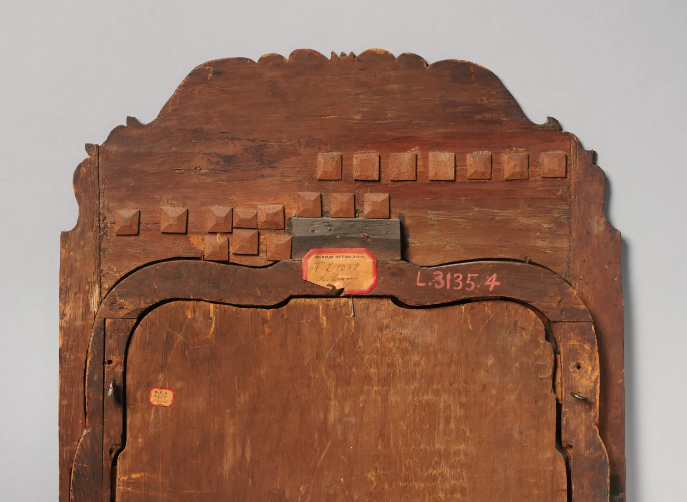 Back of the arched crest of a looking glass showing two rows of small square wooden glue blocks along the curve, an old museum label and an inventory number