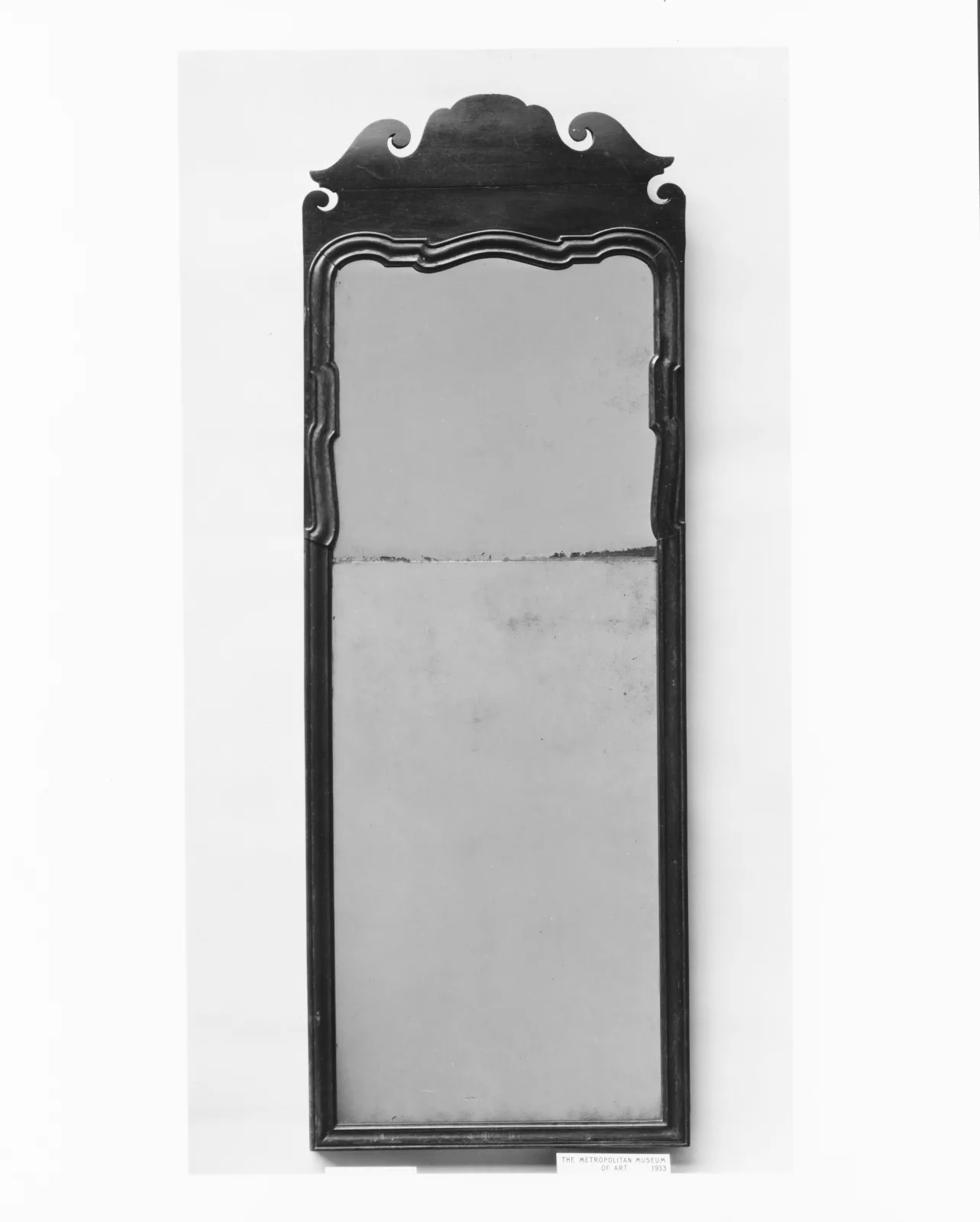 Black and white photograph of a tall walnut looking glass with a scrolled crest, the glass divided by a horizontal joint into a small upper plate and a larger lower plate