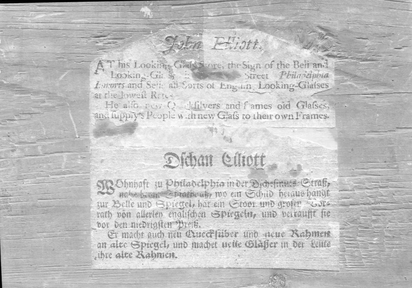 Black and white photograph of a printed paper label in English and German on a wooden backboard, headed John Elliott and Dschan Elliott
