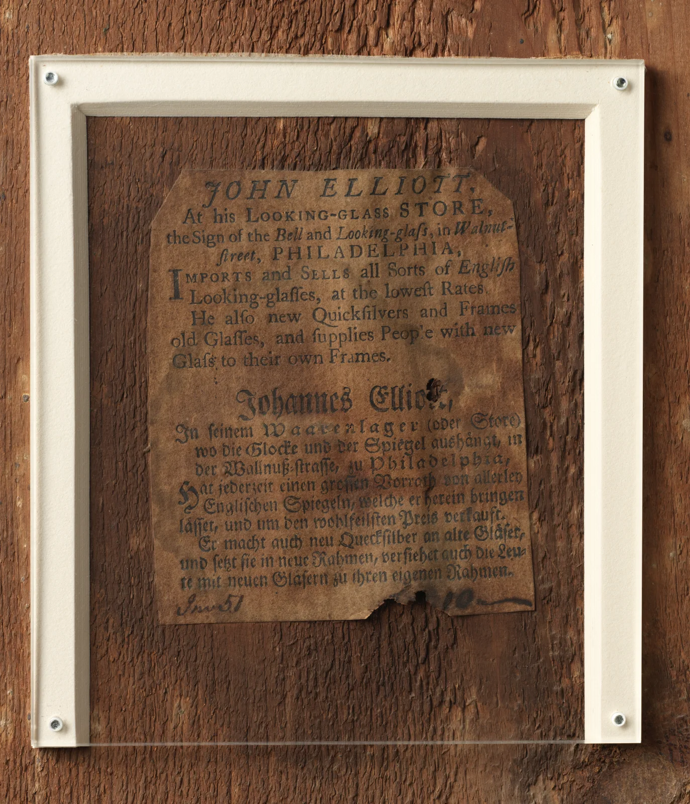 Printed paper label on a dark wooden backboard reading John Elliott, At his Looking-glass Store, the Sign of the Bell and Looking-glass, in Walnut-street, Philadelphia, with a German version below