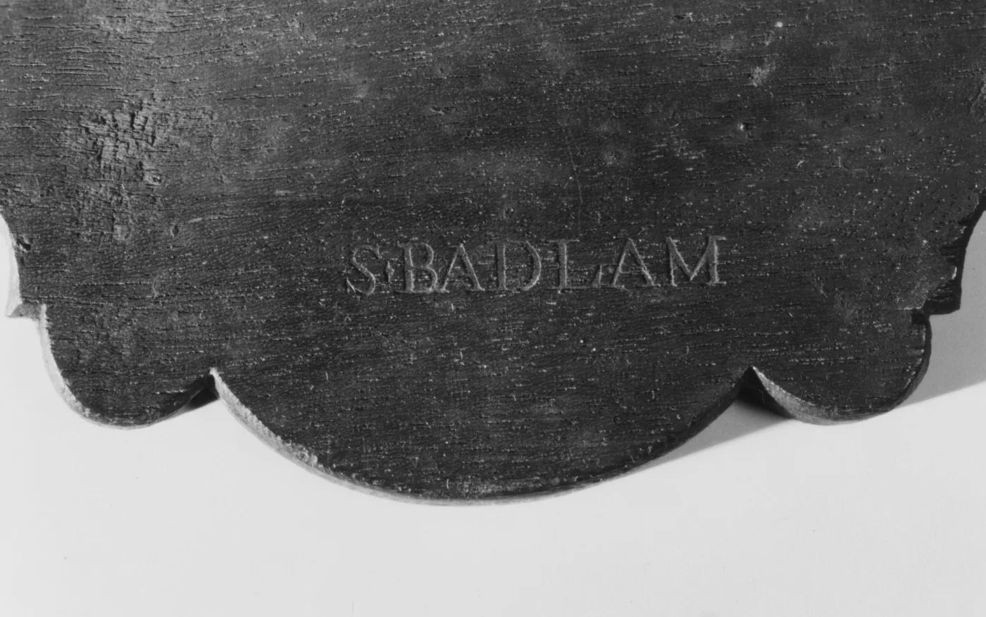 Black and white photograph of the shaped back of a looking glass crest with the name S.BADLAM stamped into the wood