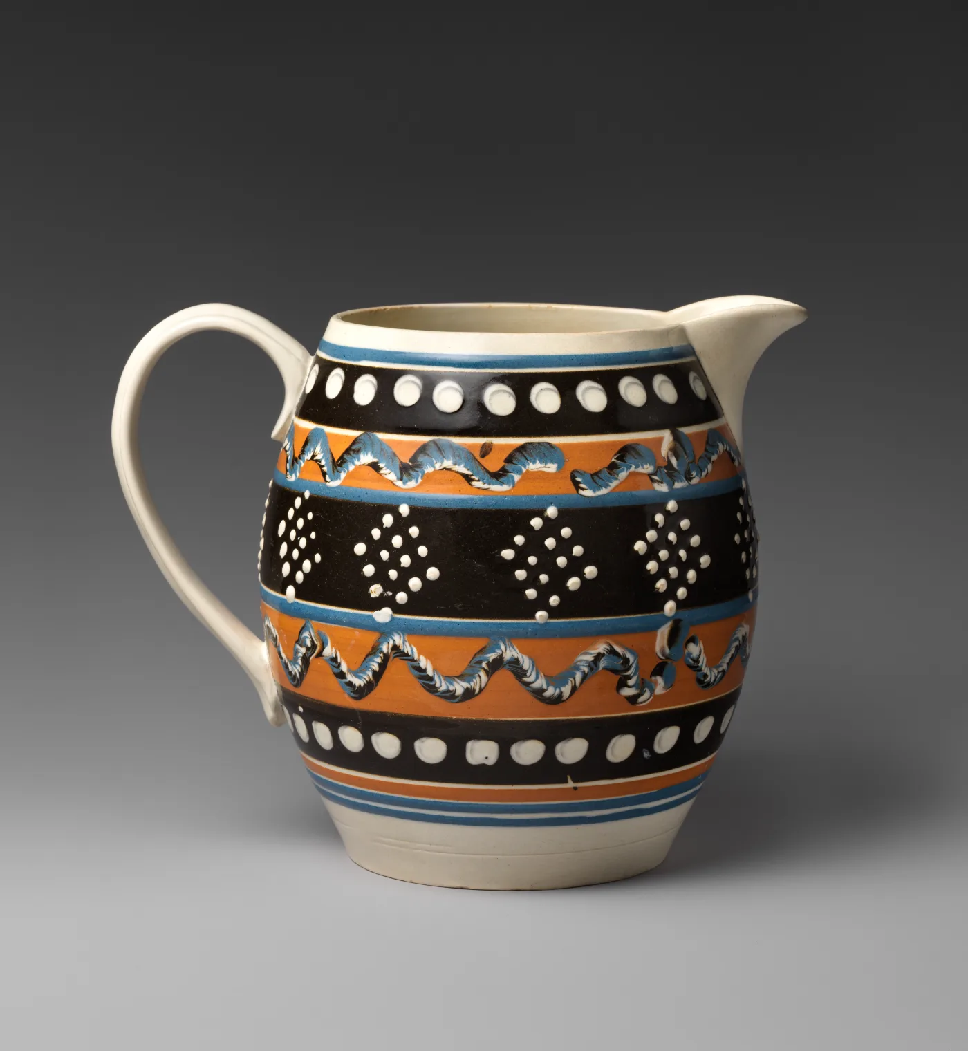 Cream-coloured jug decorated with broad horizontal bands in orange, black and blue, one band filled with rows of small ringed dots and another with a looping trailed pattern in white and brown