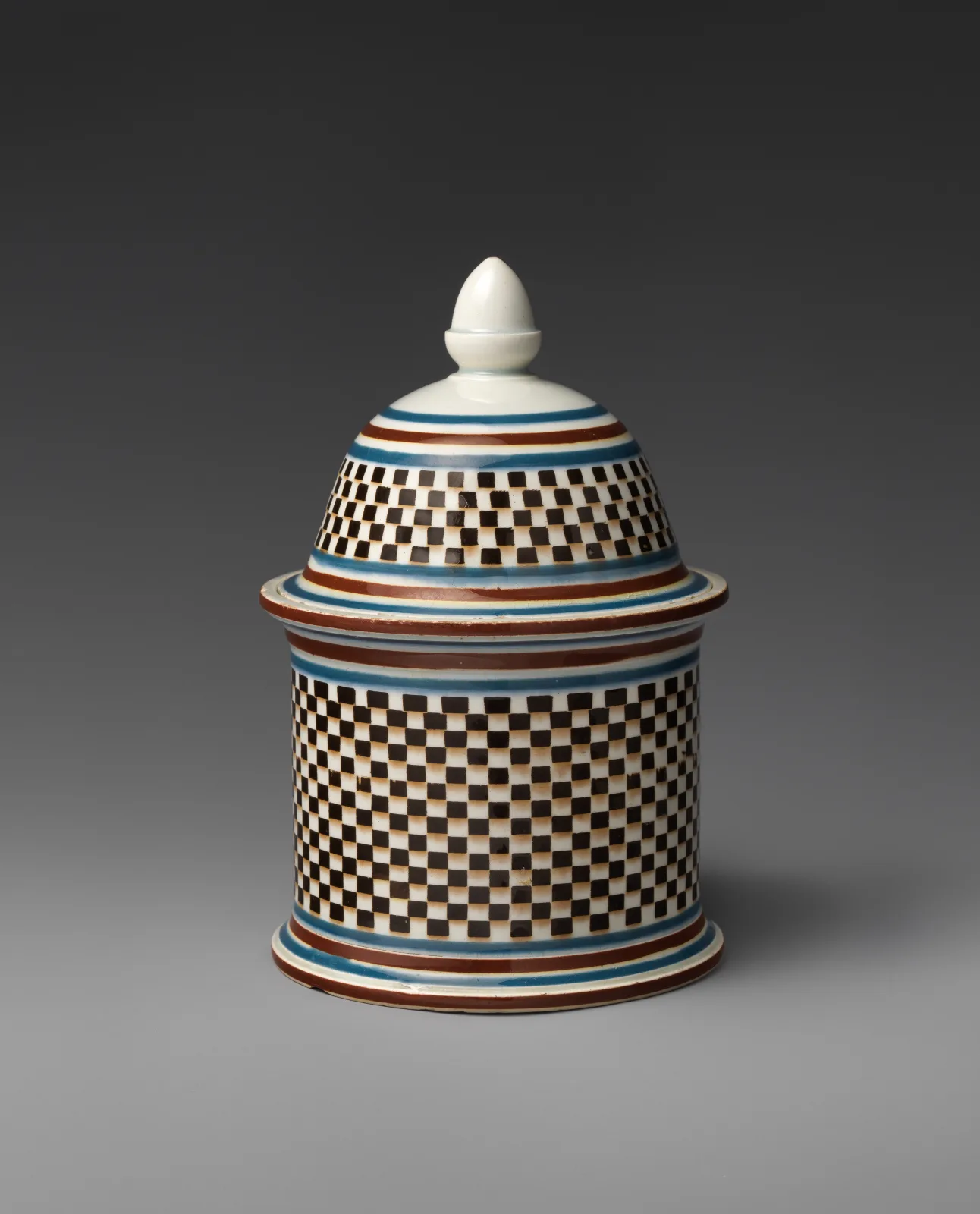 Cylindrical covered humidor decorated all over with a fine chequerboard of small squares in brown and cream, between narrow orange and blue bands