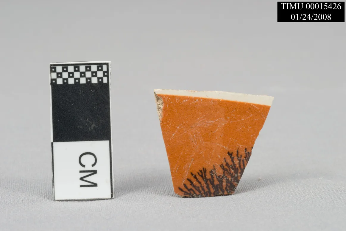 Small triangular pottery fragment with an orange slip surface carrying fine dark branching marks, photographed beside a centimetre scale bar