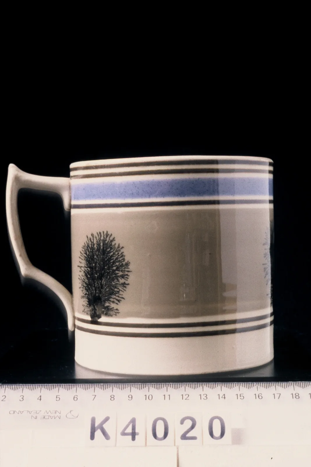 Straight-sided mug with a loop handle, a broad pale blue-grey band around the body carrying a single dark branching form like a small tree, photographed beside a centimetre rule