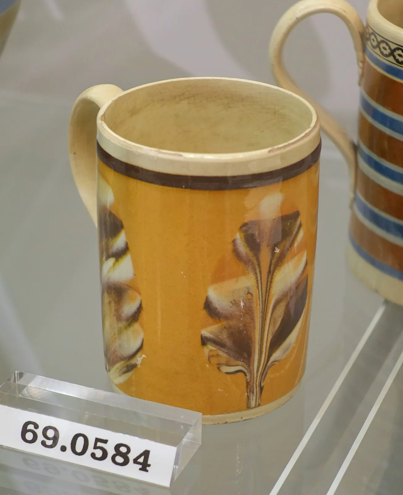 Straight-sided mug with a bright yellow ground band between dark brown borders, carrying a single dark branching dendritic form, displayed in a museum case
