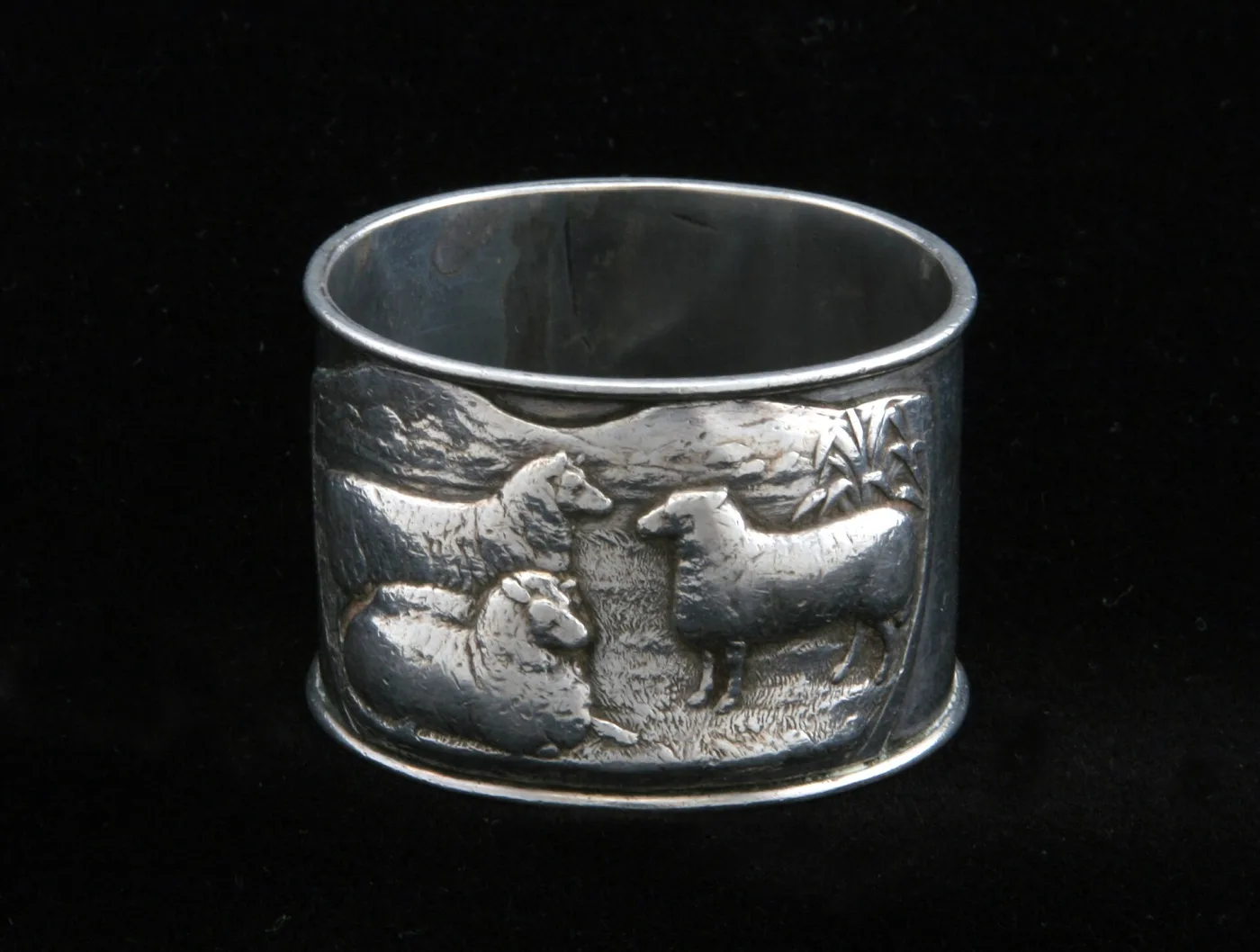 Silver napkin ring with an applied plaque showing sheep, flax and mountains in relief, photographed against black