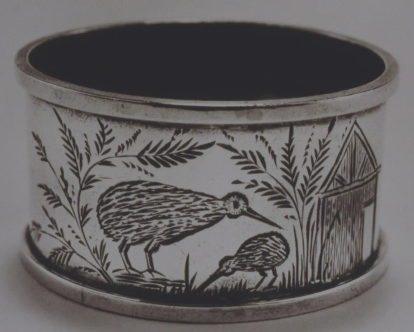 Silver napkin ring deeply engraved with two kiwi feeding among ferns beside a gable-ended wooden hut
