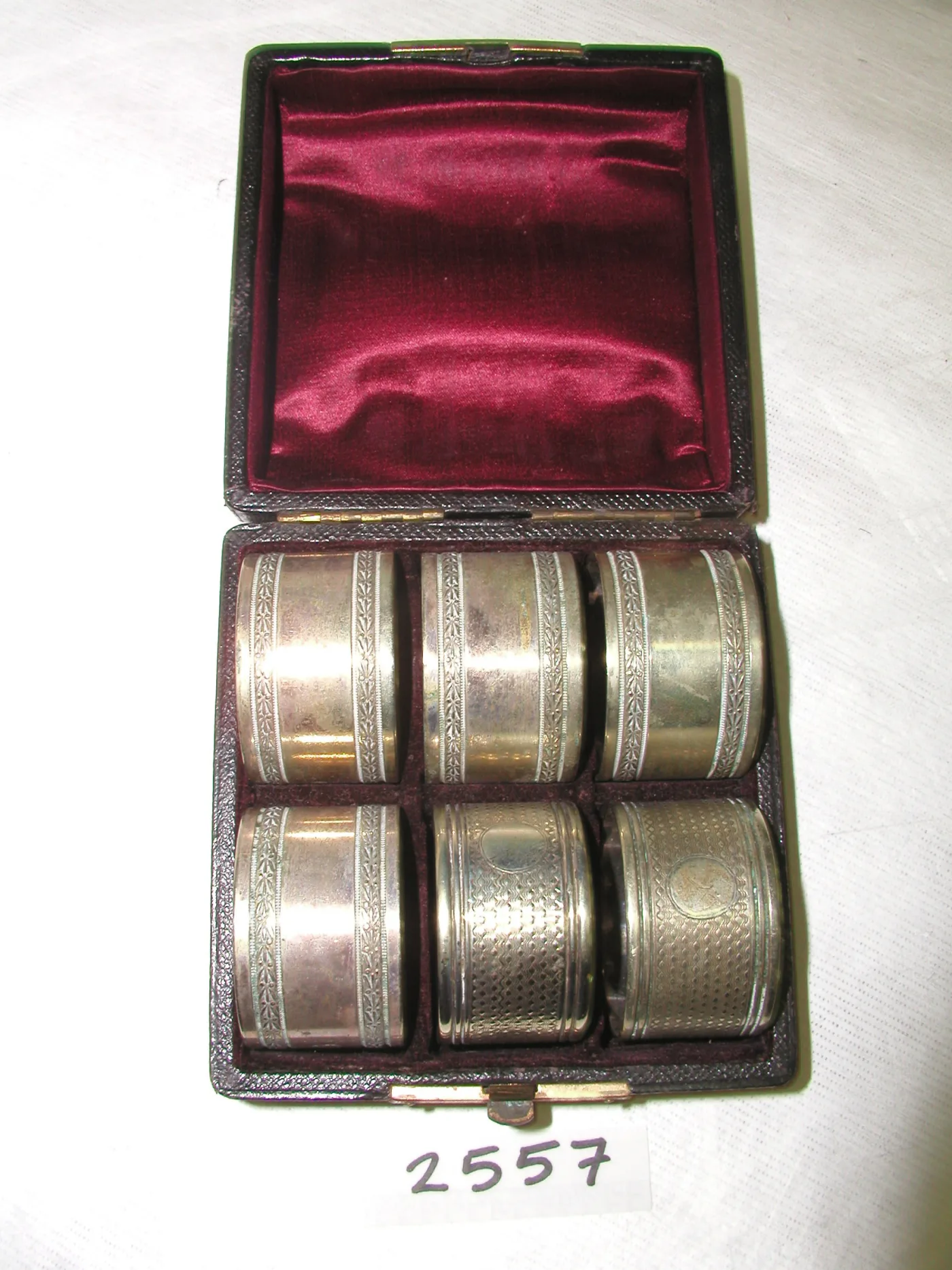 Open leather case lined with red satin holding six silver napkin rings in fitted recesses, four with one border pattern and two with another